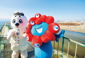 万博公式キャラクター「ミャクミャク」と建設中の万博会場をバックにツーショット